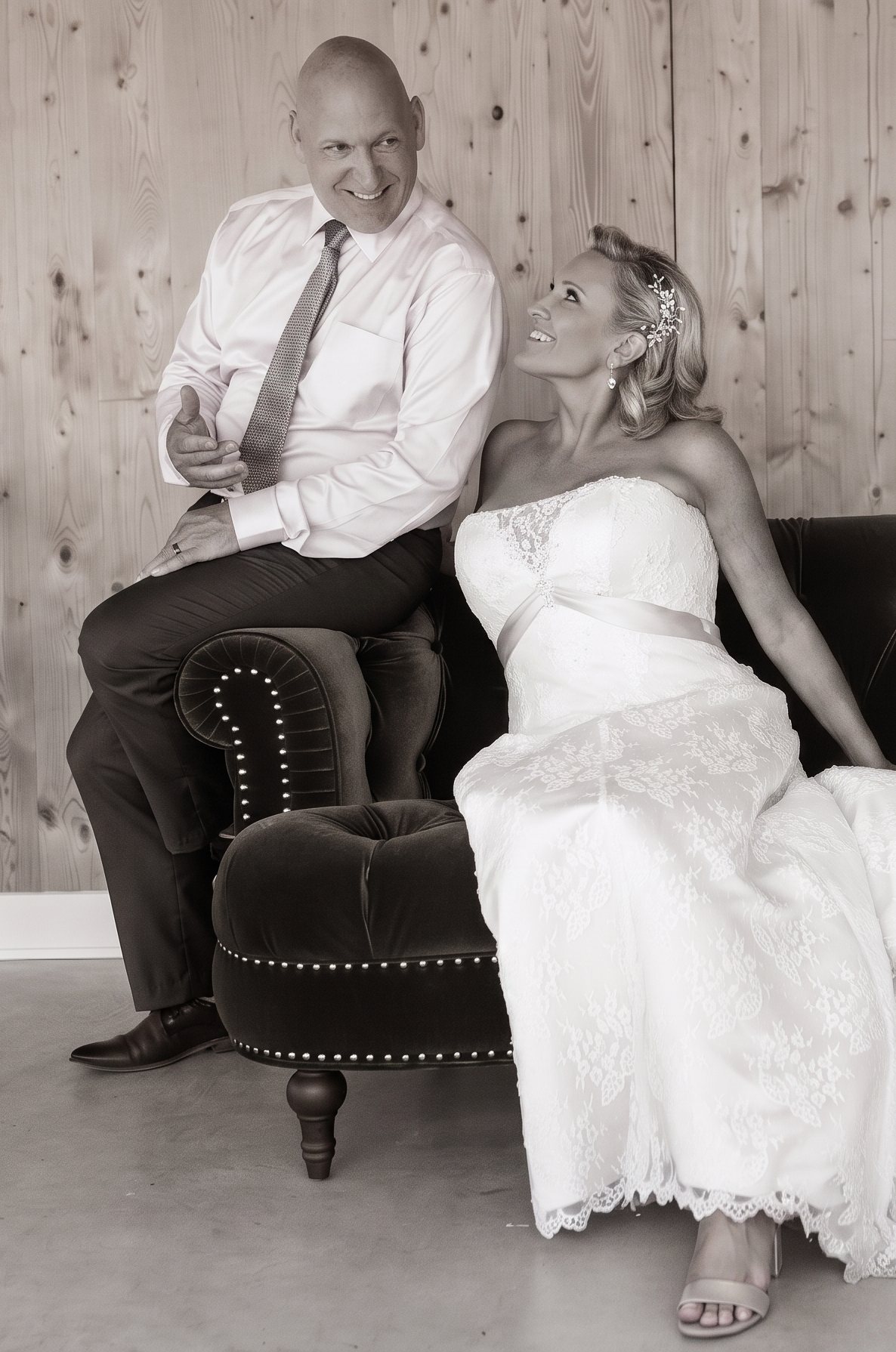 Ivan Demers and Rachelle Marquis on their wedding day in August 2025, in a sepia portrait. Ivan sits on the arm of a velvet sofa in a white shirt and tie, smiling, while Rachelle sits beside him in a lace wedding gown, smiling up at him.