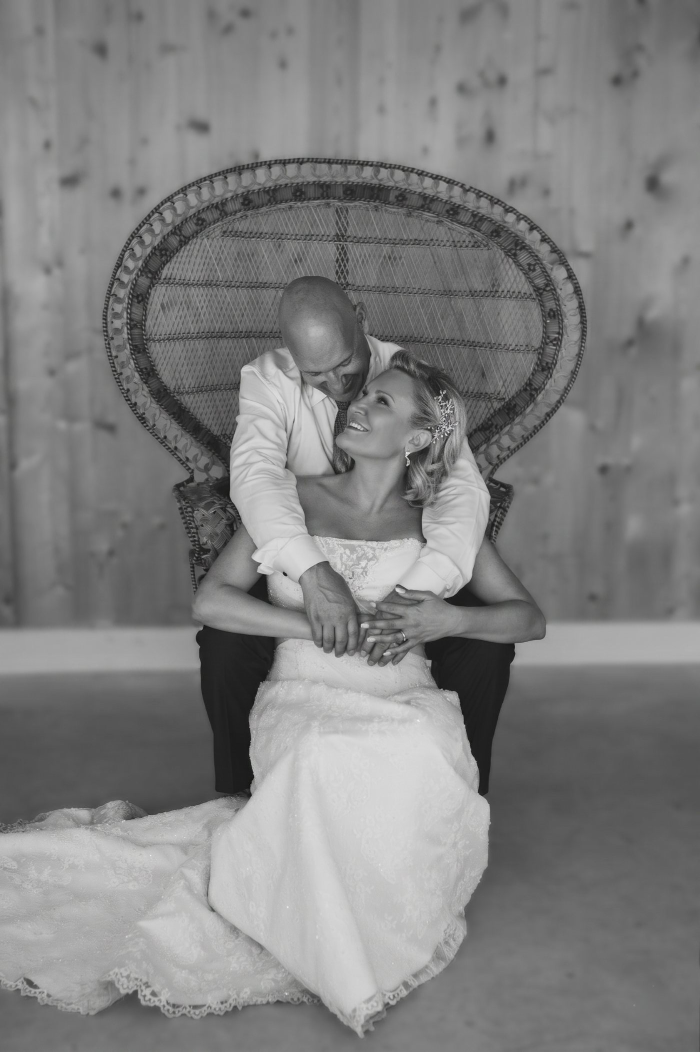 Ivan Demers and Rachelle Marquis on their wedding day in August 2025, in a black-and-white portrait. Rachelle sits in a tall wicker chair in her lace wedding gown, smiling up at Ivan, who leans over her from behind with his arms around her.